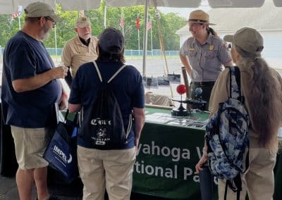 A group of people stands at a booth under a tent, speaking with two park rangers wearing uniforms. A table covered with a green cloth reads "Cuyahoga Valley National Park." Trees and flag poles are visible in the background.