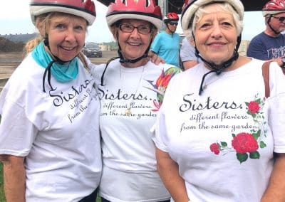 Three elderly women wearing bike helmets stand together, smiling. They wear matching shirts with a floral design and text reading, "Sisters: different flowers from the same garden."