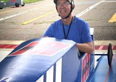 A person wearing a helmet and glasses sits smiling in a blue soapbox derby car on a paved track. The car has a number 16 and racing stripes on it. The background features grassy fields and a cloudy sky.