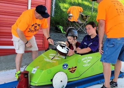Three people assist a young person in a helmet sitting in a green soapbox car. Two adults in orange shirts, one wearing a cap, are providing guidance. A folding chair and grassy hill are visible in the background.