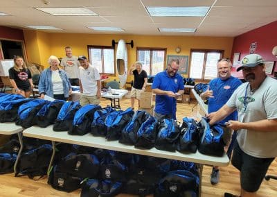 A group of people in a brightly colored room stand around tables filled with blue and black bags. They are smiling and gesturing towards the bags. Various equipment and boxes are visible in the background.