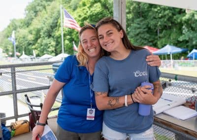 Two smiling women stand together under a pavilion, one wearing a blue shirt and lanyard, the other in a gray shirt, holding a water bottle. Flags and trees are in the background, suggesting an outdoor sports event.