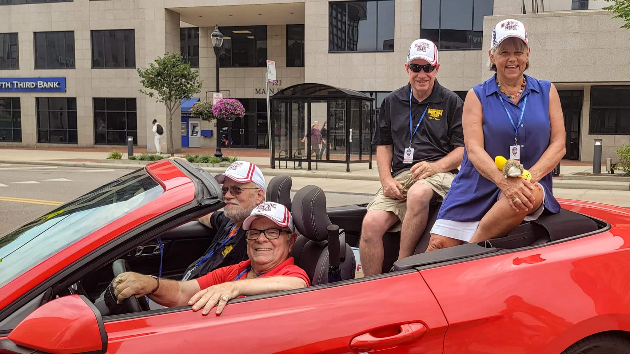 Four smiling people wearing hats and lanyards sit in and on a red convertible. The car is parked on a city street in front of a bank building.